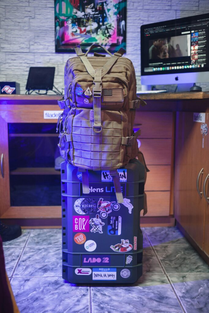Tan backpack on a sticker-covered suitcase in a sleek office with computer and decorations.